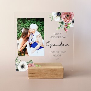 Puede incluir: Un marco de fotos de acr&iacute;lico transparente con un dise&ntilde;o floral y una foto de dos mujeres. El marco tiene el texto "Happy Mothers Day Grandma Lots of Love Felicity xx" impreso en &eacute;l. El marco est&aacute; sentado en un soporte de madera.