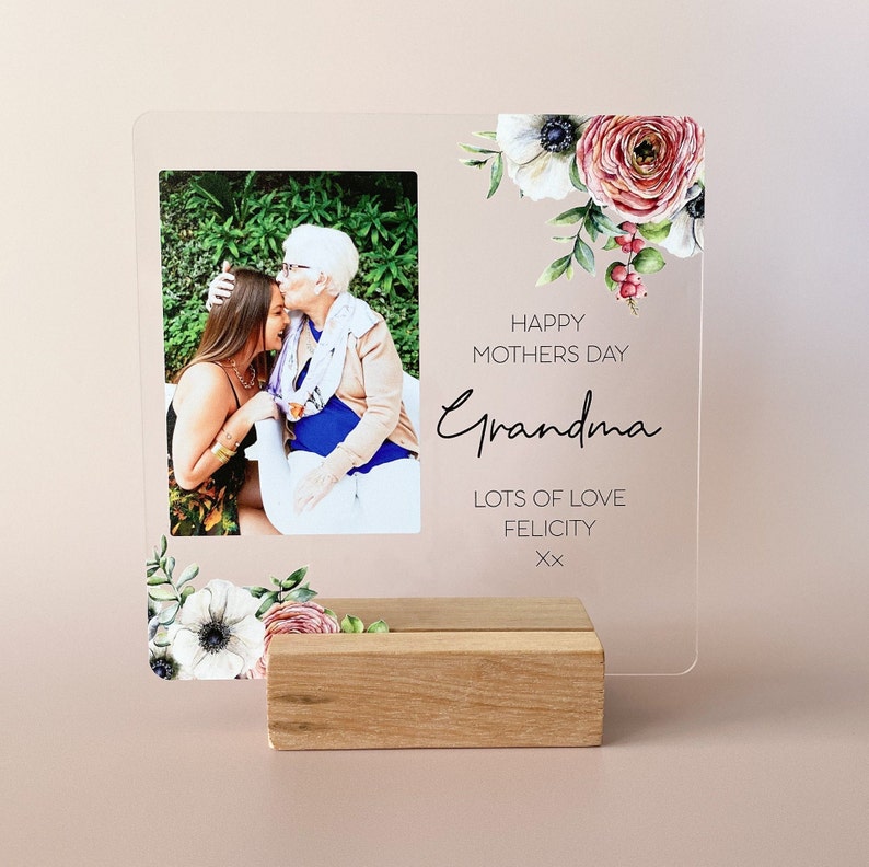 Puede incluir: Un marco de fotos de acr&iacute;lico transparente con un dise&ntilde;o floral y una foto de dos mujeres. El marco tiene el texto "Happy Mothers Day Grandma Lots of Love Felicity xx" impreso en &eacute;l. El marco est&aacute; sentado en un soporte de madera.