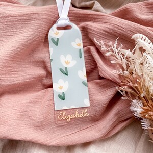 May include: A light blue bookmark with a white floral pattern and the name "Elizabeth" written in gold lettering. The bookmark is attached to a white ribbon.