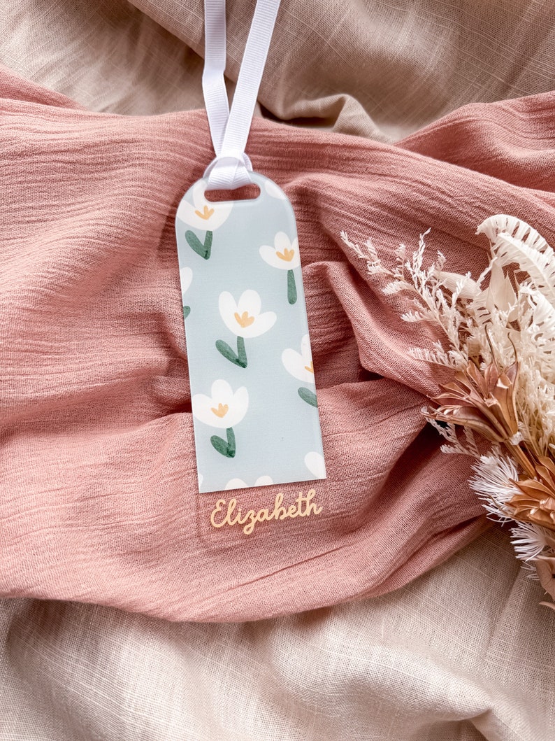 May include: A light blue bookmark with a white floral pattern and the name "Elizabeth" written in gold lettering. The bookmark is attached to a white ribbon.