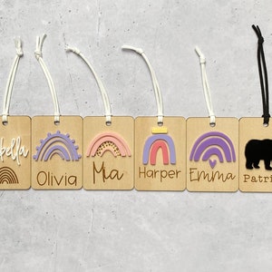 Puede incluir: Ocho etiquetas de madera con dise&ntilde;os de arco&iacute;ris coloridos y nombres como Charlotte, Isabella, Olivia, Mia, Harper, Emma, Patrick y Archie. Las etiquetas est&aacute;n unidas a cordones blancos y negros.