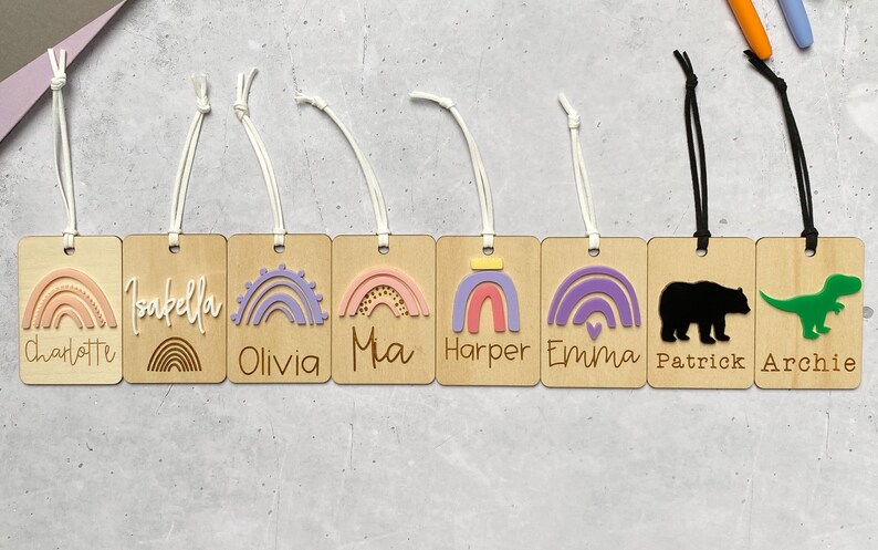 Puede incluir: Ocho etiquetas de madera con dise&ntilde;os de arco&iacute;ris coloridos y nombres como Charlotte, Isabella, Olivia, Mia, Harper, Emma, Patrick y Archie. Las etiquetas est&aacute;n unidas a cordones blancos y negros.
