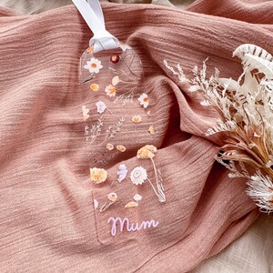 May include: A clear bookmark with pressed flowers and greenery. The bookmark is shaped like a long rectangle with rounded corners. The word "Mum" is written in pink lettering at the bottom of the bookmark.