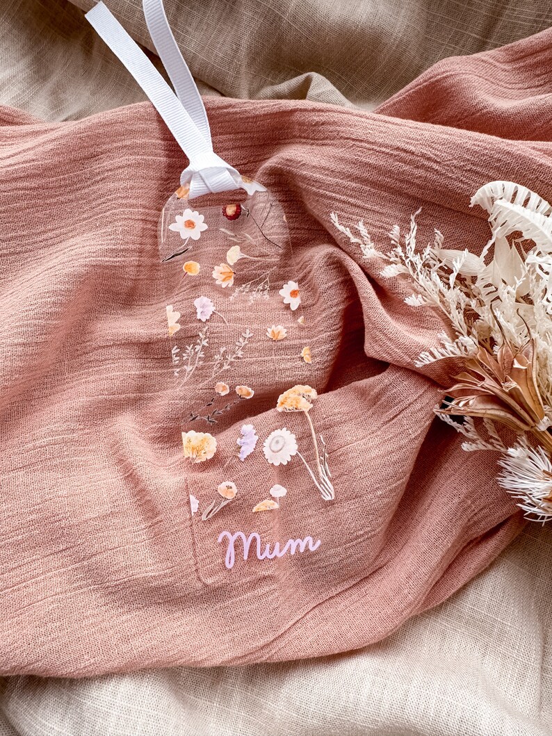 May include: A clear bookmark with pressed flowers and greenery. The bookmark is shaped like a long rectangle with rounded corners. The word "Mum" is written in pink lettering at the bottom of the bookmark.