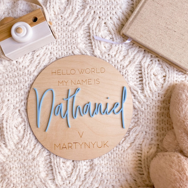 May include: A circular wooden plaque featuring the engraved text HELLO WORLD MY NAME IS and MARTYNYUK, with the name Nathaniel in raised, light blue script lettering centered on the surface of the light-colored wood material.