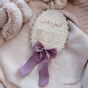 May include: An oval wooden birth announcement sign with a scalloped edge, featuring the engraved text Hello World and lines for Name, Date, Time, Weight, and Length. A dusty purple fabric bow is attached to the bottom of the light wood plaque.