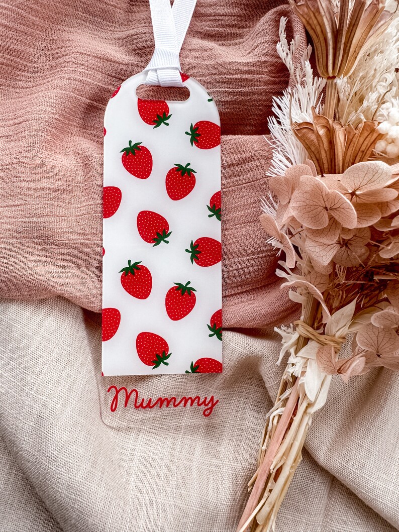 May include: A white bookmark with a red strawberry pattern and the word "Mummy" written in red on the bottom. The bookmark has a white ribbon attached to the top.