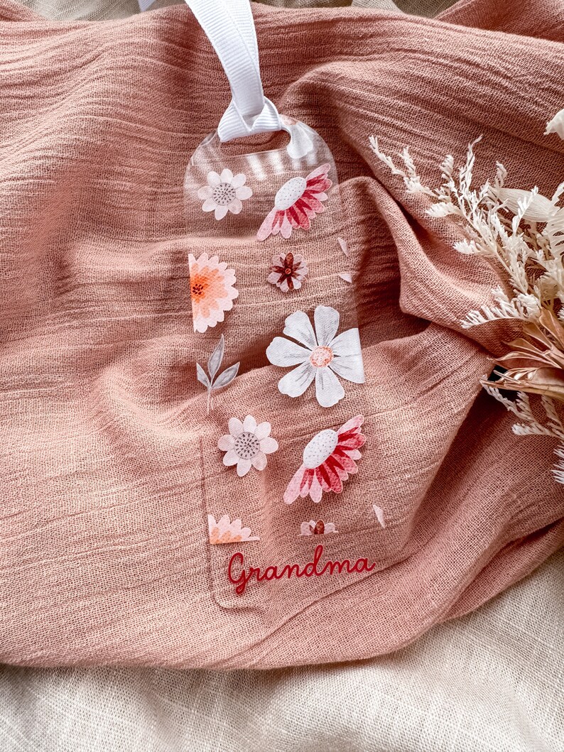 May include: A clear acrylic bookmark with a floral design and the word "Grandma" in red lettering. The bookmark is hanging from a white ribbon.