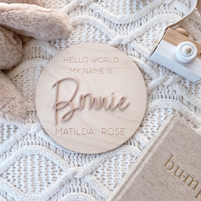 May include: A circular light-colored wooden plaque featuring the engraved text HELLO WORLD MY NAME IS and MATILDA ROSE, with the name Bonnie in raised, script-style lettering centered on the surface of the wood disc.