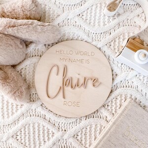 May include: A circular light-colored wooden plaque featuring the engraved text HELLO WORLD MY NAME IS and ROSE, with the name Claire in raised, script-style lettering centered on the surface. The plaque is positioned among soft textures.