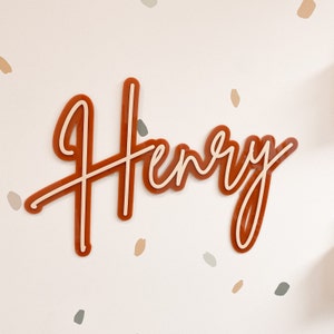 May include: A custom name sign with the name "Henry" in a cursive font. The sign is made of a thick, orange-brown acrylic material with a white outline.