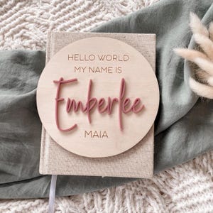 May include: A circular wooden plaque featuring the engraved text HELLO WORLD MY NAME IS and MAIA, with the name Emberlee in raised, dusty rose-colored script lettering. The plaque is centered on a beige fabric-covered book with a textured surface.