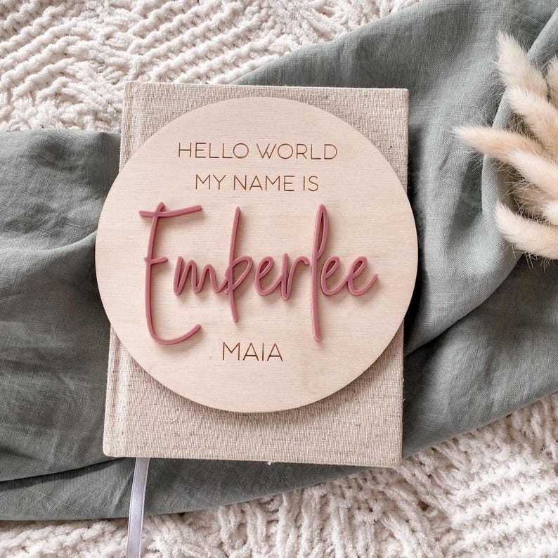 May include: A circular wooden plaque featuring the engraved text HELLO WORLD MY NAME IS and MAIA, with the name Emberlee in raised, dusty rose-colored script lettering. The plaque is centered on a beige fabric-covered book with a textured surface.