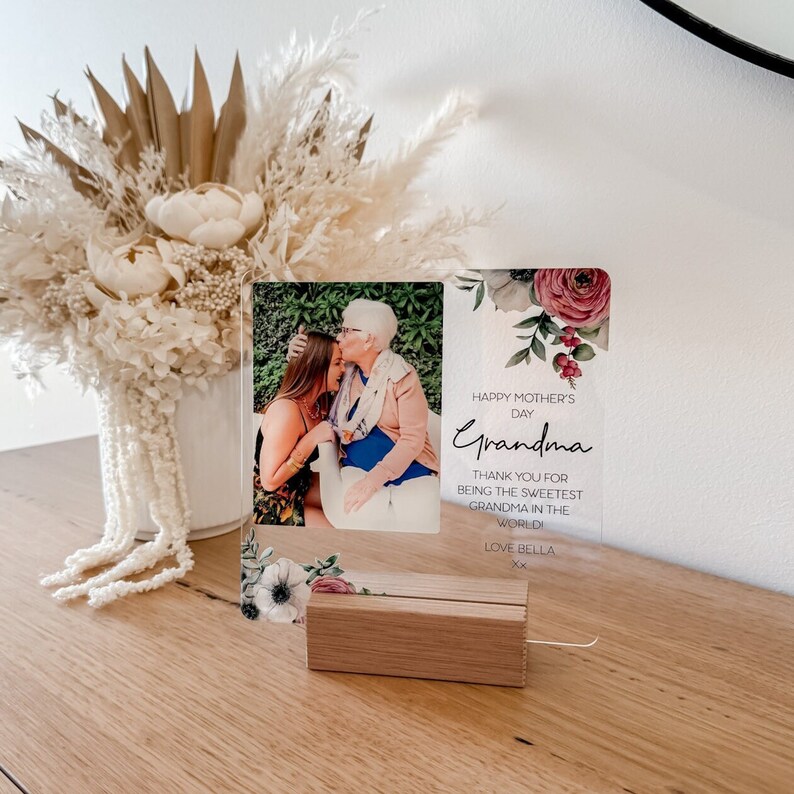 Puede incluir: Un marco de fotos de acr&iacute;lico transparente con un dise&ntilde;o floral y una foto de dos personas. El marco tiene un soporte de madera y el texto "Happy Mother's Day Grandma Thank you for being the sweetest grandma in the world Love Bella xx" est&aacute; impreso en el marco.