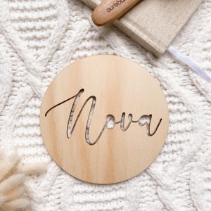Op de afbeelding: Een rond houten bord met de naam "Nova" uitgesneden in een cursief schrift.