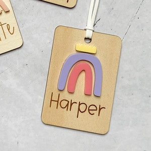 Puede incluir: Una etiqueta de madera con un dise&ntilde;o de arco&iacute;ris y el nombre "Harper" grabado en ella. El arco&iacute;ris est&aacute; hecho de acr&iacute;lico rosa, morado y amarillo.