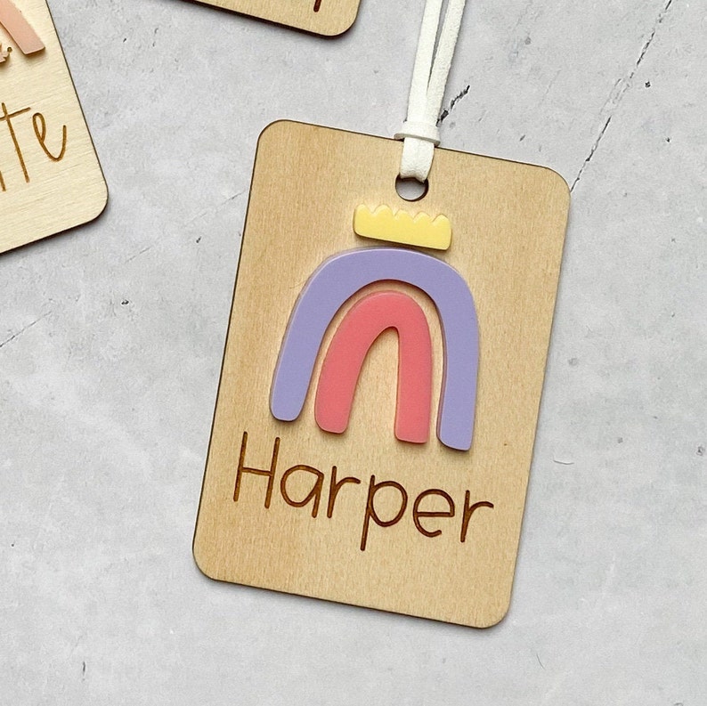 Puede incluir: Una etiqueta de madera con un dise&ntilde;o de arco&iacute;ris y el nombre "Harper" grabado en ella. El arco&iacute;ris est&aacute; hecho de acr&iacute;lico rosa, morado y amarillo.