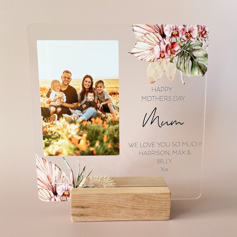 Puede incluir: Un marco de fotos de acr&iacute;lico transparente con un dise&ntilde;o floral y una foto de familia. El marco tiene una base de madera y el texto "Happy Mothers Day Mum We love you so much! Harrison, Max & Billy xx" est&aacute; impreso en el marco.