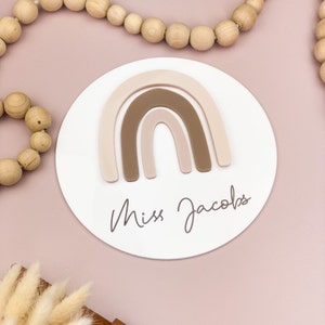 May include: A white round plaque with a brown and pink rainbow design and the text "Miss Jacobs" written in cursive.