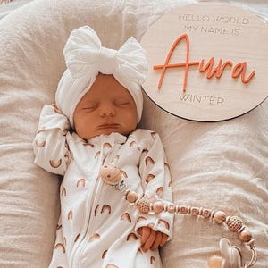 May include: A newborn wearing a white turban with a large bow and a white onesie featuring a repeating rainbow pattern. Beside the infant is a round wooden sign with the text Hello World My Name Is Aura Winter and a wooden pacifier clip.