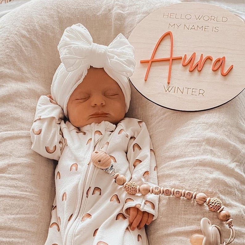 May include: A newborn wearing a white turban with a large bow and a white onesie featuring a repeating rainbow pattern. Beside the infant is a round wooden sign with the text Hello World My Name Is Aura Winter and a wooden pacifier clip.