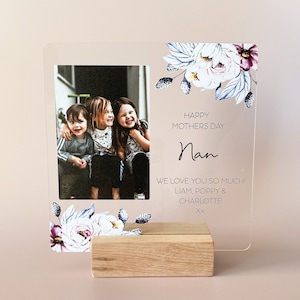Puede incluir: Un marco de fotos de acr&iacute;lico transparente con un dise&ntilde;o floral y una foto de tres ni&ntilde;os. El marco tiene una base de madera y el texto "Happy Mothers Day Nan We love you so much! Liam, Poppy & Charlotte Xx" est&aacute; impreso en el marco.