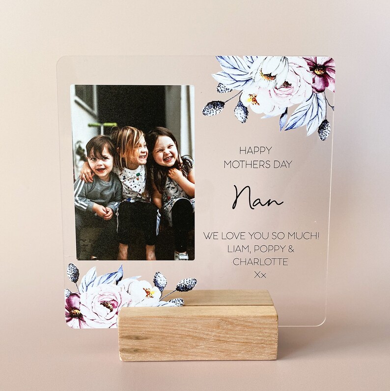 Puede incluir: Un marco de fotos de acr&iacute;lico transparente con un dise&ntilde;o floral y una foto de tres ni&ntilde;os. El marco tiene una base de madera y el texto "Happy Mothers Day Nan We love you so much! Liam, Poppy & Charlotte Xx" est&aacute; impreso en el marco.