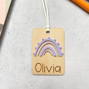 Puede incluir: Una etiqueta de madera con un dise&ntilde;o de arco&iacute;ris morado y el nombre "Olivia" grabado en ella. La etiqueta est&aacute; unida a una cuerda blanca.