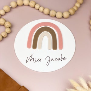 May include: White round plaque with a pink, brown, and beige rainbow design. The text "Miss Jacobs" is written in cursive below the rainbow.