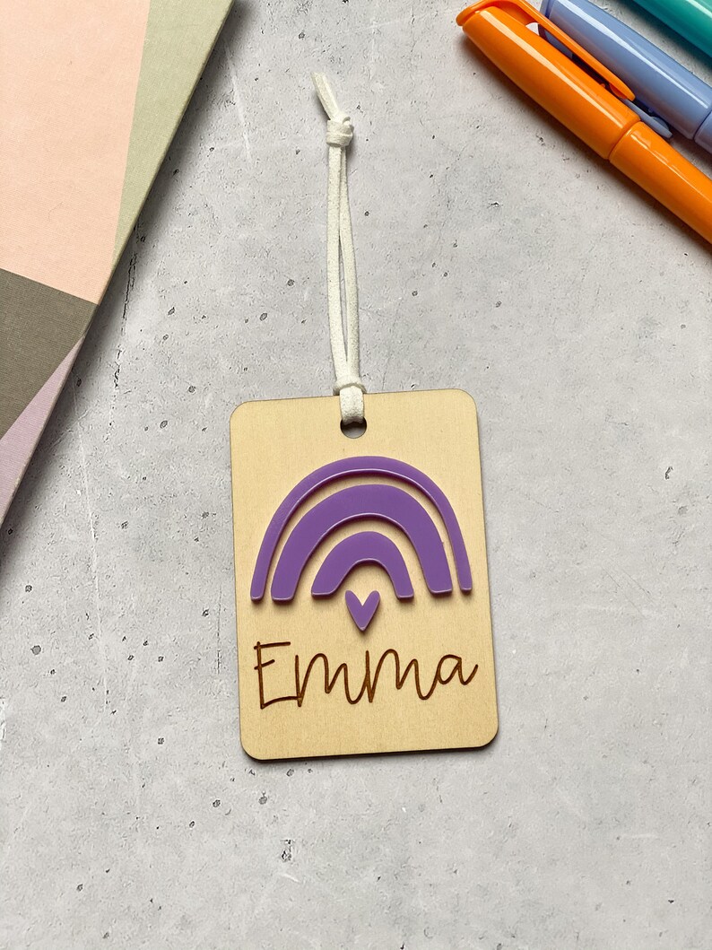 Puede incluir: Una etiqueta de madera con un arco&iacute;ris morado y el nombre "Emma" grabado en ella. La etiqueta tiene una cuerda blanca atada.