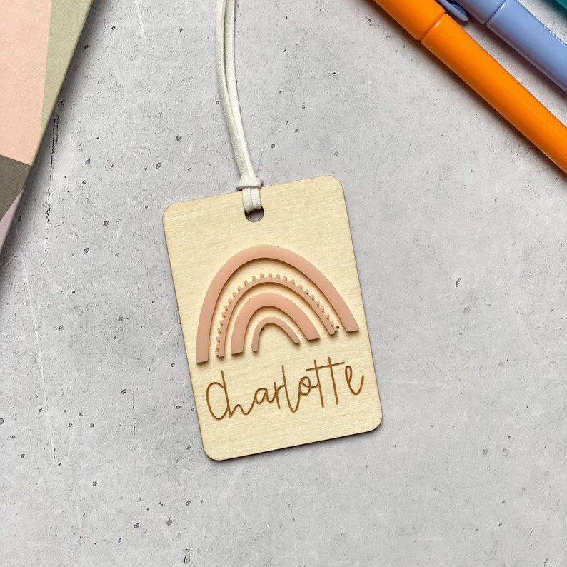Puede incluir: Una etiqueta de madera con un dise&ntilde;o de arco&iacute;ris rosa y el nombre "Charlotte" grabado en letras marrones. La etiqueta tiene una cuerda blanca.