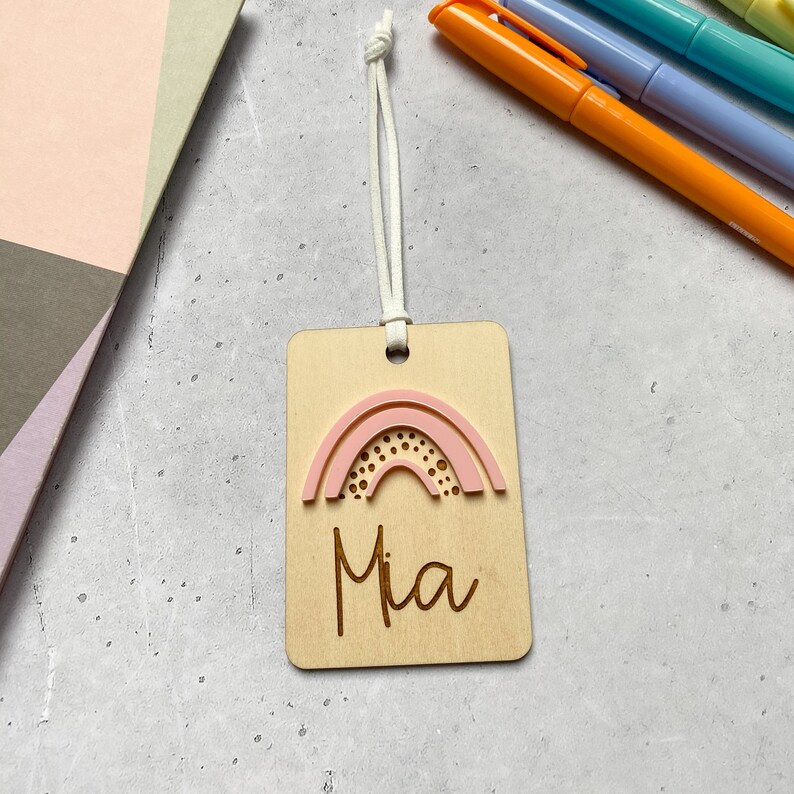 Puede incluir: Una etiqueta de madera con un dise&ntilde;o de arco&iacute;ris rosa y el nombre "Mia" grabado en ella. La etiqueta est&aacute; unida a una cuerda blanca.
