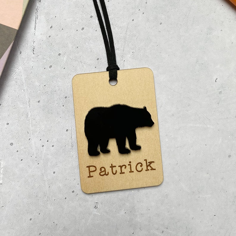 Puede incluir: Una etiqueta de madera con una silueta de oso negro y el nombre "Patrick" grabado en ella. La etiqueta est&aacute; unida a un cord&oacute;n negro.