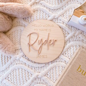 May include: A circular wooden plaque featuring the engraved text HELLO WORLD MY NAME IS and JAMES, with the name Ryder in raised, script-style lettering centered on the surface. The plaque has a light wood grain finish and smooth edges.