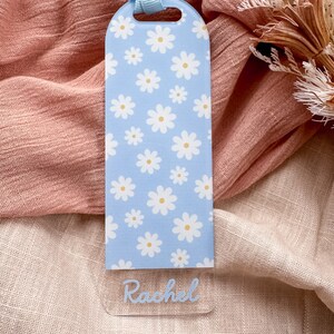 May include: A light blue bookmark with a white daisy pattern and the name "Rachel" written in blue script.