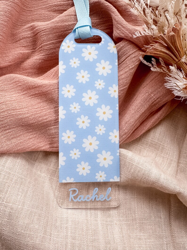 May include: A light blue bookmark with a white daisy pattern and the name "Rachel" written in blue script.