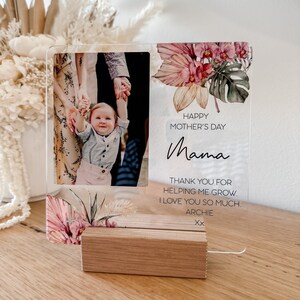Puede incluir: Un marco de fotos de acr&iacute;lico transparente con un dise&ntilde;o floral y una foto de una familia. El texto "Happy Mother's Day Mama" est&aacute; impreso en el marco. El texto "Thank you for helping me grow. I love you so much. Archie Xx" tambi&eacute;n est&aacute; impreso en el marco.