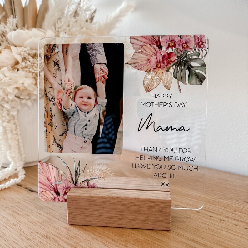 Puede incluir: Un marco de fotos de acr&iacute;lico transparente con un dise&ntilde;o floral y una foto de una familia. El texto "Happy Mother's Day Mama" est&aacute; impreso en el marco. El texto "Thank you for helping me grow. I love you so much. Archie Xx" tambi&eacute;n est&aacute; impreso en el marco.
