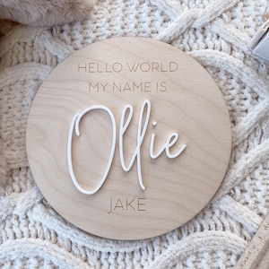 May include: A circular light-colored wooden plaque featuring the engraved text HELLO WORLD MY NAME IS and JAKE. The name Ollie is displayed in the center in raised white 3D script lettering, creating a layered effect on the smooth wood surface.
