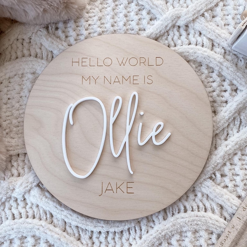 May include: A circular light-colored wooden plaque featuring the engraved text HELLO WORLD MY NAME IS and JAKE. The name Ollie is displayed in the center in raised white 3D script lettering, creating a layered effect on the smooth wood surface.