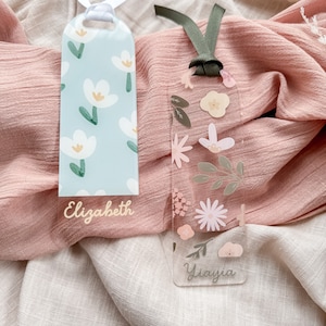 May include: Two personalized bookmarks with floral designs. The bookmark on the left is light blue with white flowers and the text "Elizabeth". The bookmark on the right is clear with pink and green flowers and the text "Yiavjia".