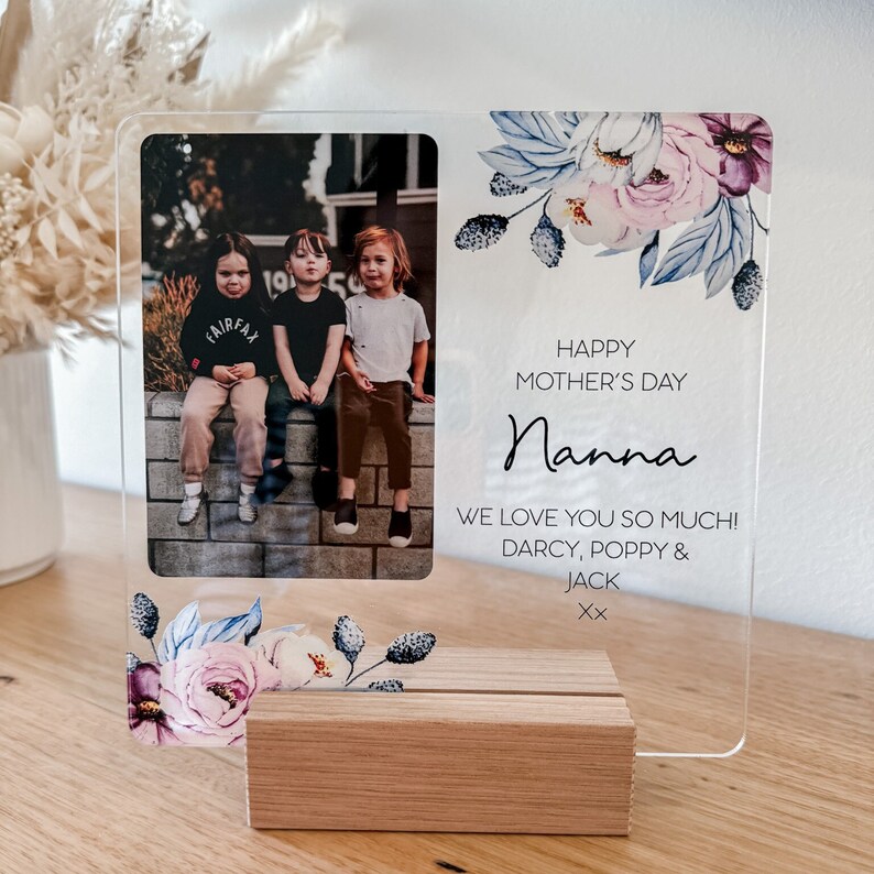 Puede incluir: Un marco de fotos de acr&iacute;lico transparente con un dise&ntilde;o floral y una foto de tres ni&ntilde;os. El marco tiene un soporte de madera y el texto "Happy Mother's Day Nanna We love you so much! Darcy, Poppy & Jack Xx" est&aacute; impreso en el marco.