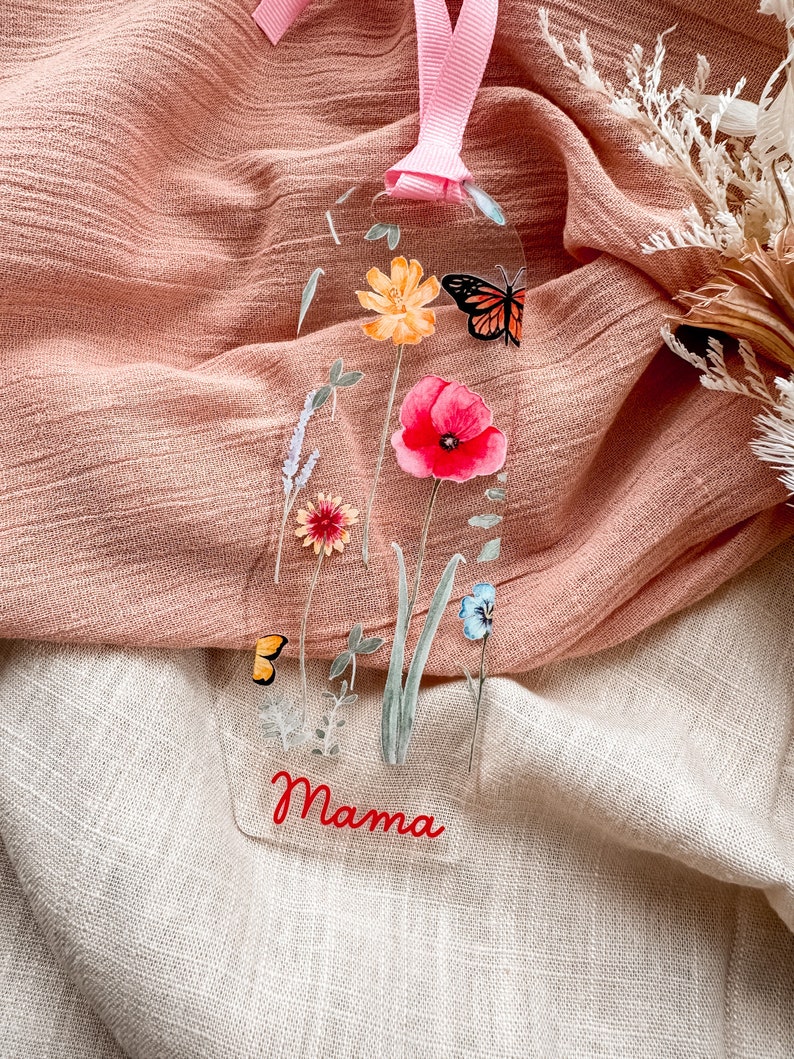 May include: A clear bookmark with a floral design featuring a butterfly, poppy, and other flowers. The bookmark has the word "Mama" written in red at the bottom.
