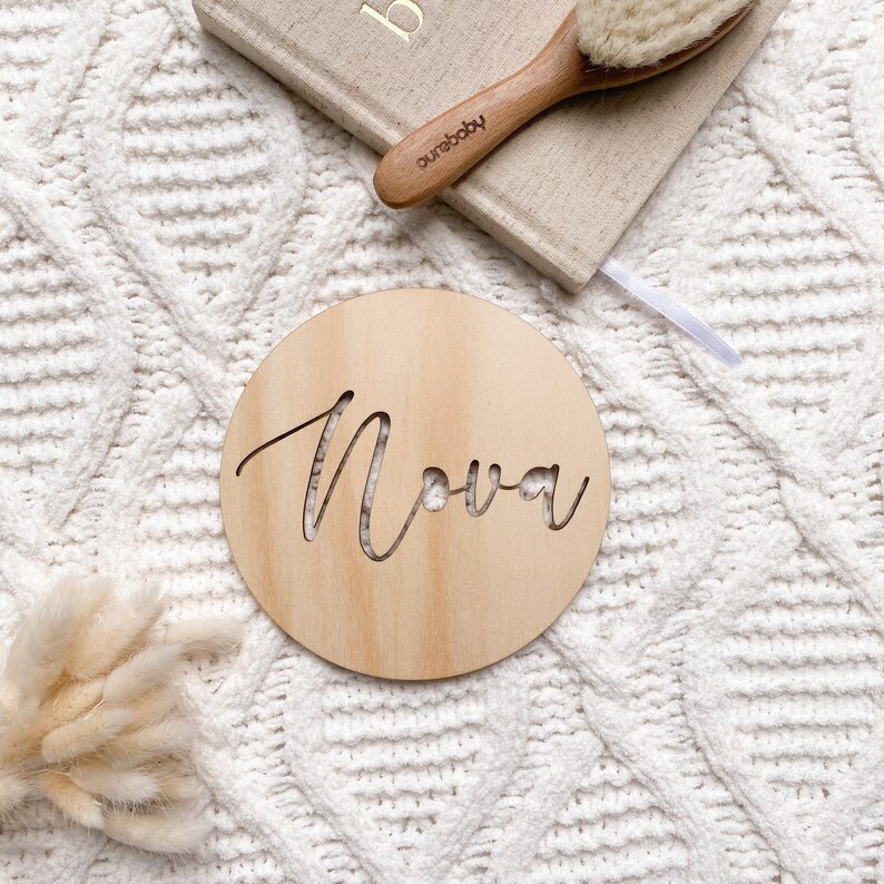 Op de afbeelding: Een ronde houten plaquette met de naam "Nova" erin gegraveerd in een cursief schrift. De plaquette is lichtbruin en heeft een natuurlijke houten afwerking.