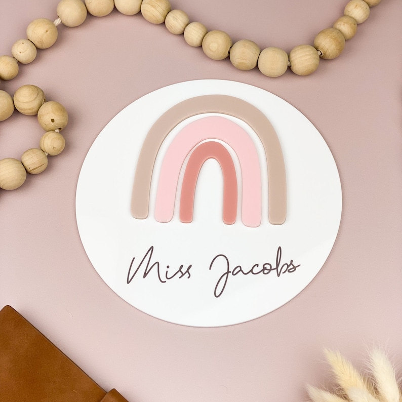 May include: A white round sign with a pink and brown rainbow design and the text "Miss Jacobs" in cursive.