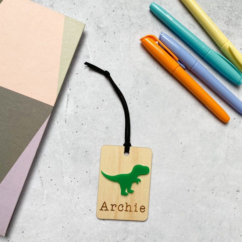 Puede incluir: Un marcap&aacute;ginas de madera con una silueta de dinosaurio verde y el nombre "Archie" grabado en &eacute;l. El marcap&aacute;ginas est&aacute; unido a una cuerda negra.