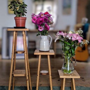 May include: Three wooden plant stands of varying heights. The stands hold a potted plant, a vase of pink flowers, and a glass vase of lilies. The stands are made of light-coloured wood and have a simple, minimalist design.