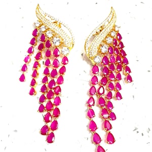 May include: A pair of gold-toned earrings with a cascading design. Each earring features a curved, leaf-like top encrusted with small, clear stones. Below, rows of vibrant, teardrop-shaped, fuchsia-colored stones dangle, creating a glamorous effect.