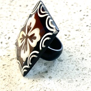 May include: A square-shaped ring with a brown and white floral design. The ring is made of resin and has a black band.