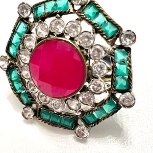 May include: A ring featuring a large, round, pink stone surrounded by smaller, clear stones and green stones in a geometric design.